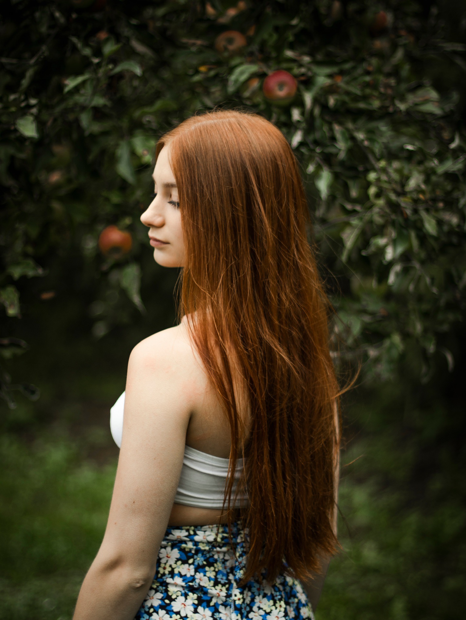 Portret z profilu kobiety z długimi rudymi włosami, ubrana w biały top i kwiecistą spódnicę, na tle drzewa jabłoni z czerwonymi owocami.