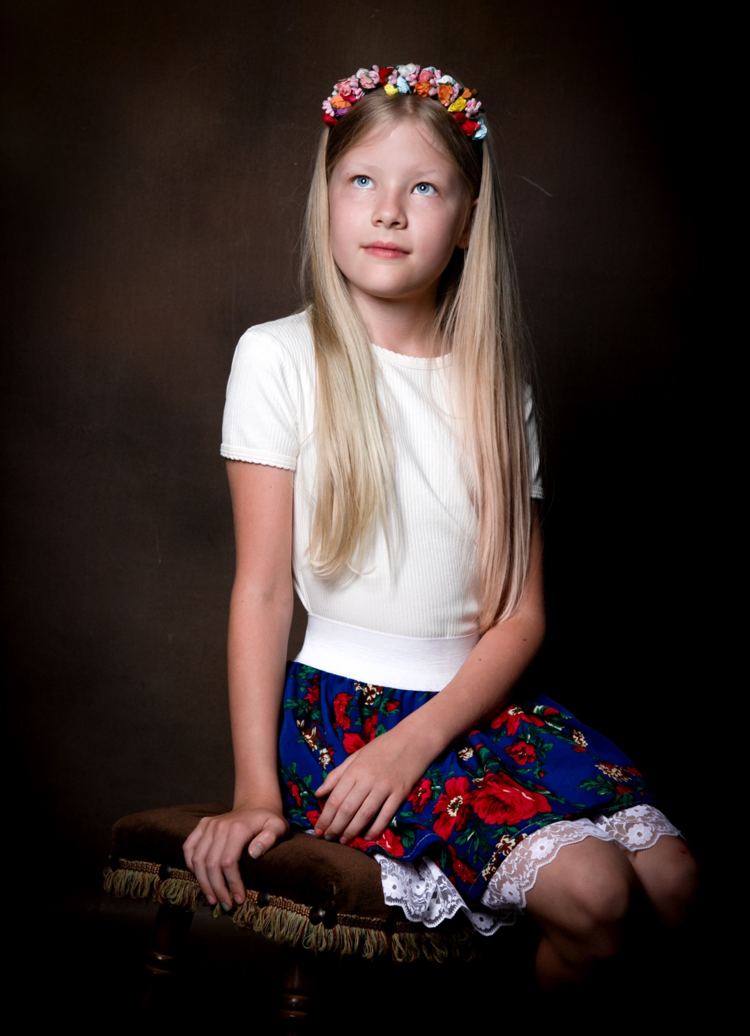 Portret dziewczynki z blond włosami i wiankiem z kwiatów na głowie, siedzącej na brązowym taborecie na ciemnym tle. Ubrana w białą bluzkę i kwiecistą spódnicę.