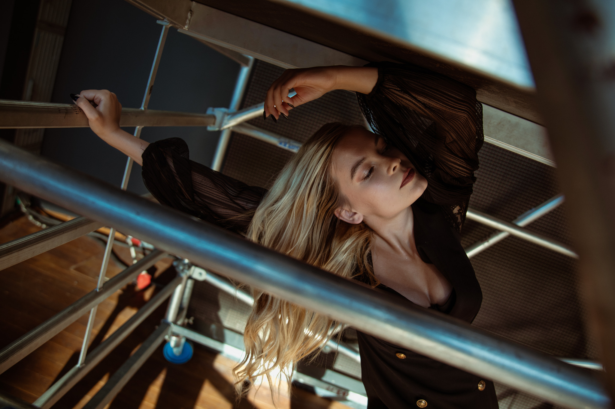 Artystyczne ujęcie kobiety w czarnej bluzce z bufiastymi rękawami, leżącej na metalowej konstrukcji rusztowania, z zamkniętymi oczami i rozpuszczonymi blond włosami.