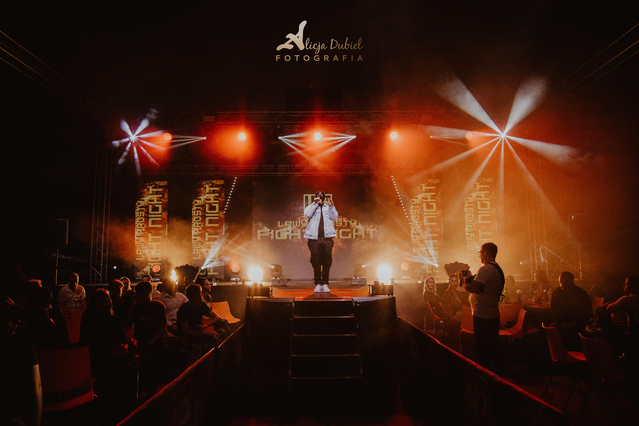 Piosenkarz na scenie podczas koncertu 'Lewy Prosty Fight Night', oświetlony reflektorami, z widownią w tle i logo fotografa w górnej części kadru.