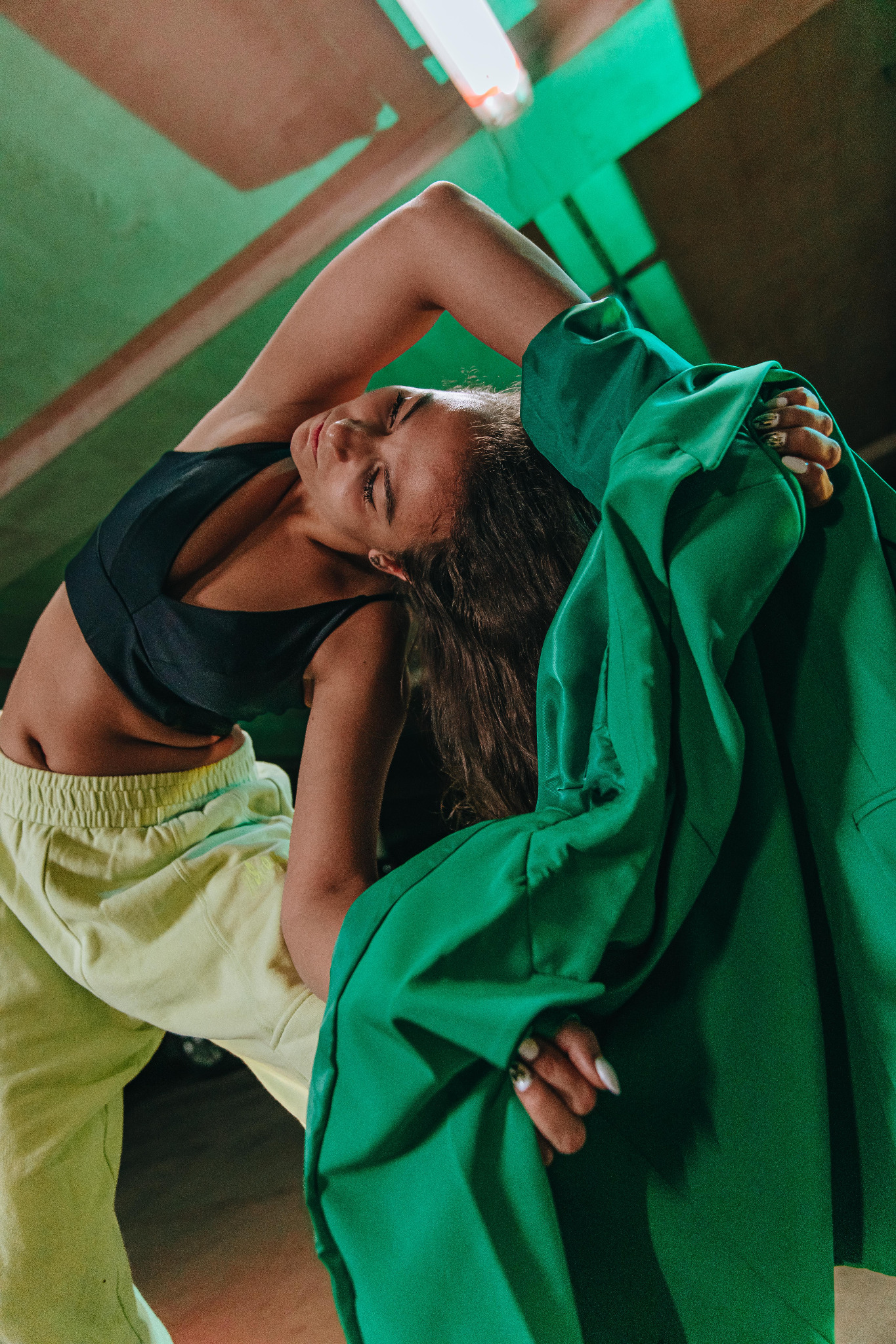 Kobieta w sportowym topie i luźnych spodniach, z zieloną marynarką, wygina się pod światłem lampy w garażu, prezentując stylizację.