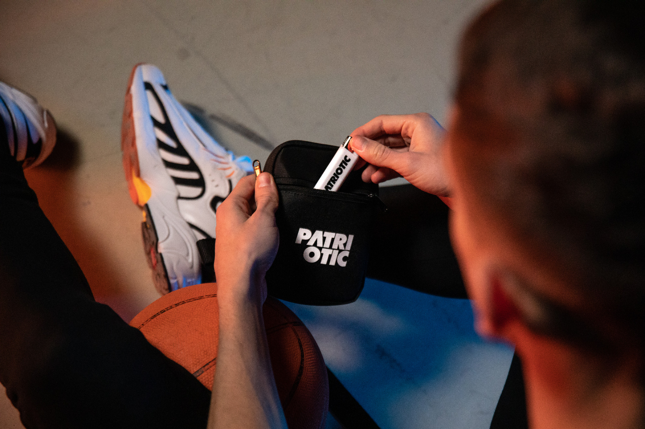 Mężczyzna wyjmuje z czarnej saszetki z napisem 'PATRIOTIC' biały długopis, obok leżą sportowe buty i piłka do koszykówki.