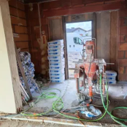 Wnętrze w trakcie prac wykończeniowych z maszyną do tynkowania, workami z zaprawą Knauf MP 75 Diamant, widoczne przewody i elementy konstrukcyjne ściany z cegły dziurawki.