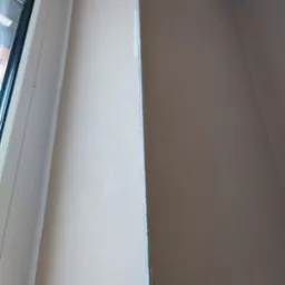 Świeżo nałożona, jasna powłoka na ścianie w rogu pomieszczenia, widoczna listwa narożna przy oknie, sugerująca etap wykończeniowy.