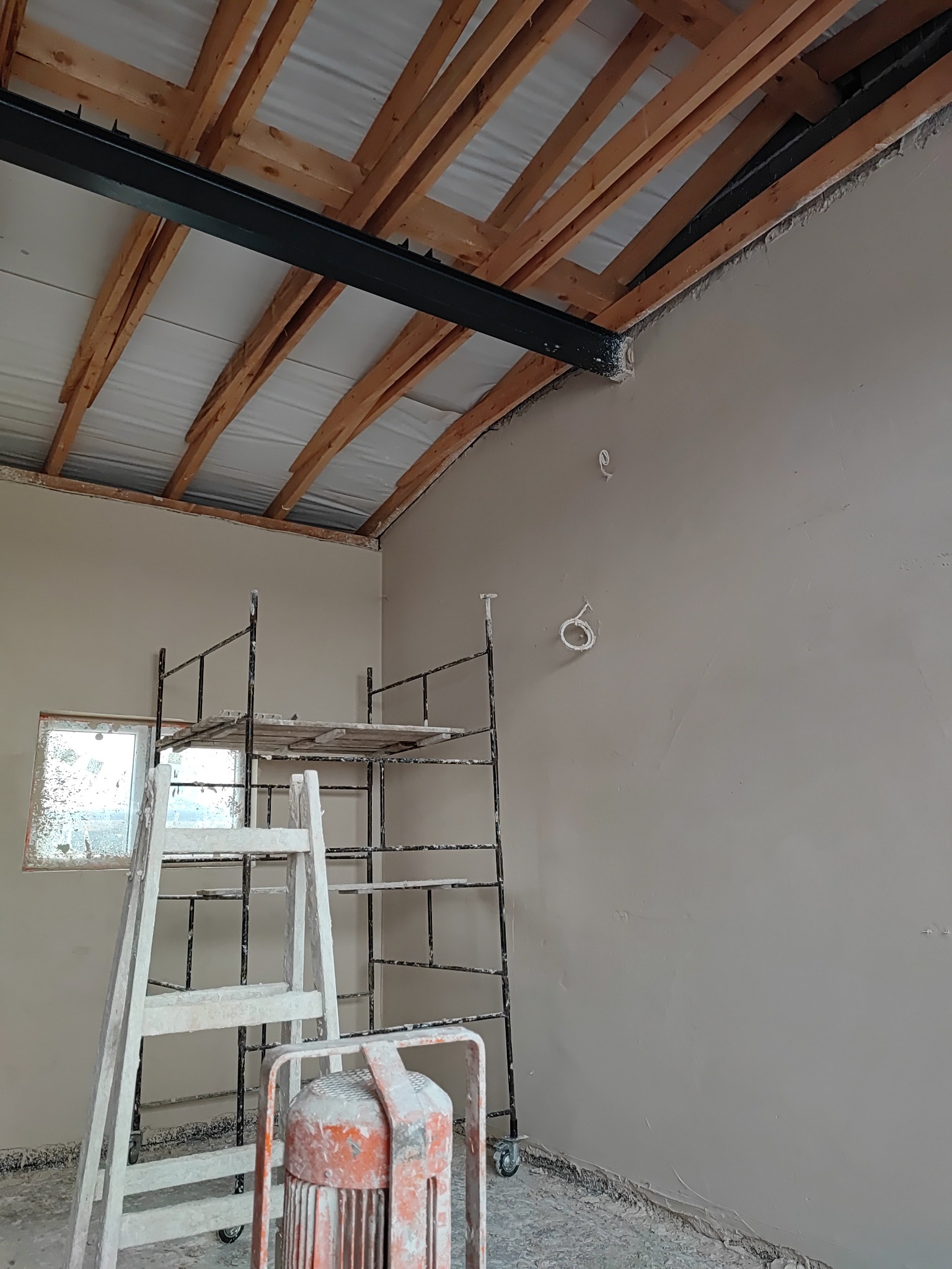 Wnętrze w trakcie tynkowania maszynowego: świeżo otynkowana ściana, metalowy ruszt z drewnianym podestem, drabina i nagrzewnica w pomieszczeniu z drewnianą konstrukcją dachu i metalową belką.