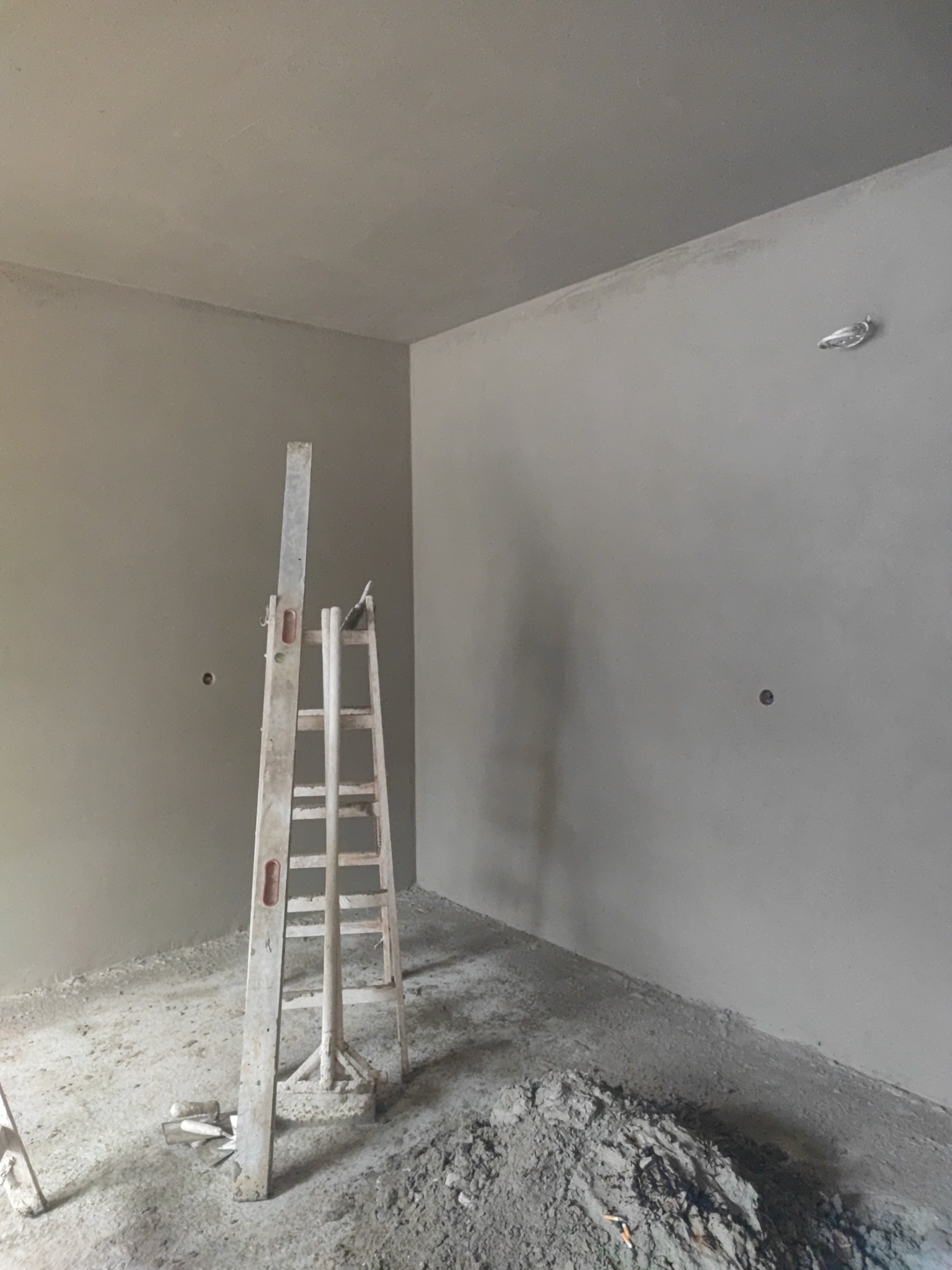 Surowe wnętrze po nałożeniu tynku, widoczna drewniana drabina, narzędzia i pozostałości materiału na podłodze, przygotowane do dalszych prac wykończeniowych.