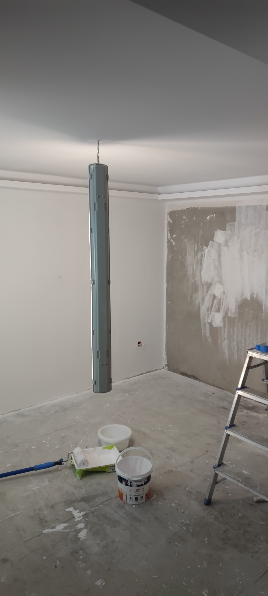 Wnętrze w trakcie malowania: surowa ściana z fragmentami gładzi, wisząca pionowa lampa budowlana, wiadro z farbą, wałek malarski i drabina.