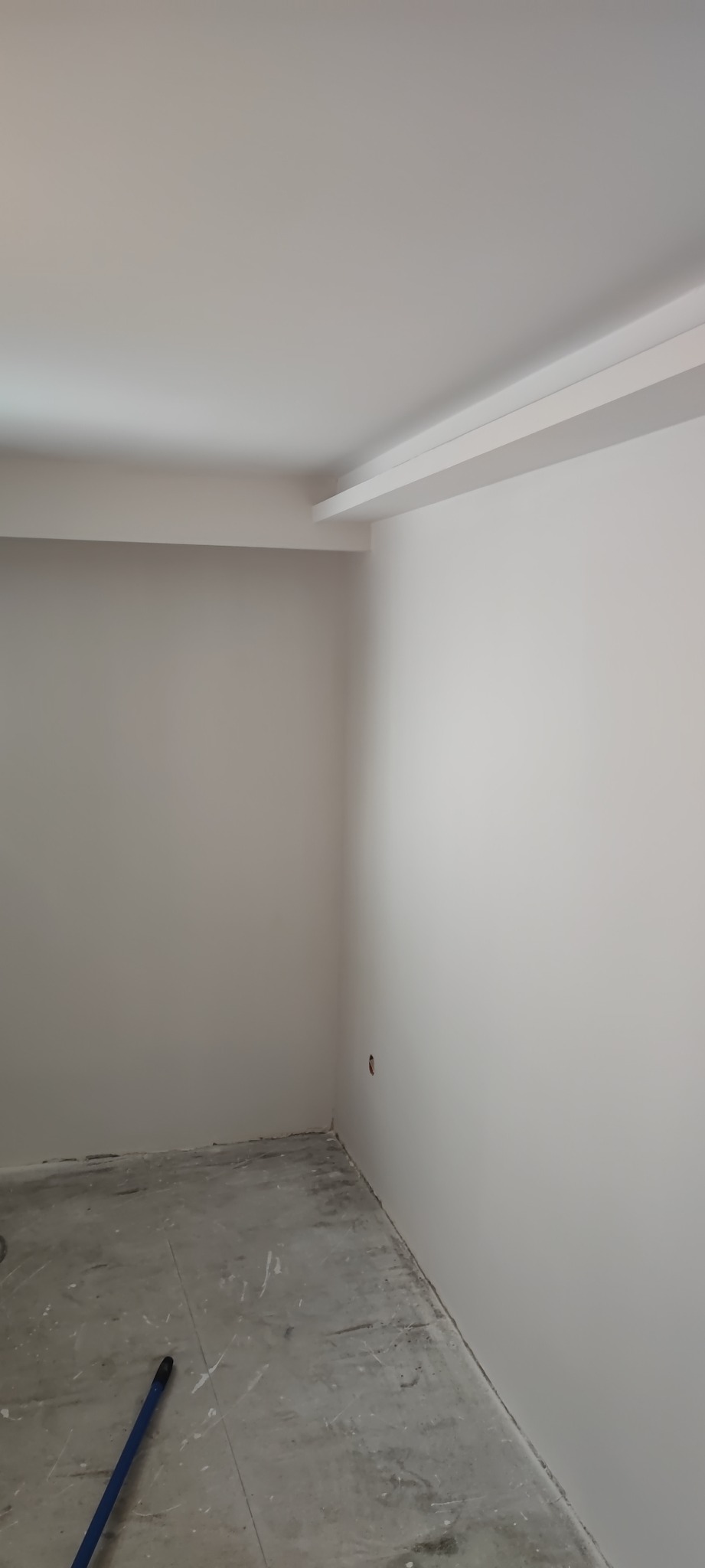 Wnętrze pokoju po malowaniu ścian na biało, widoczne ślady farby na betonowej podłodze i niebieski kij od szczotki w rogu.