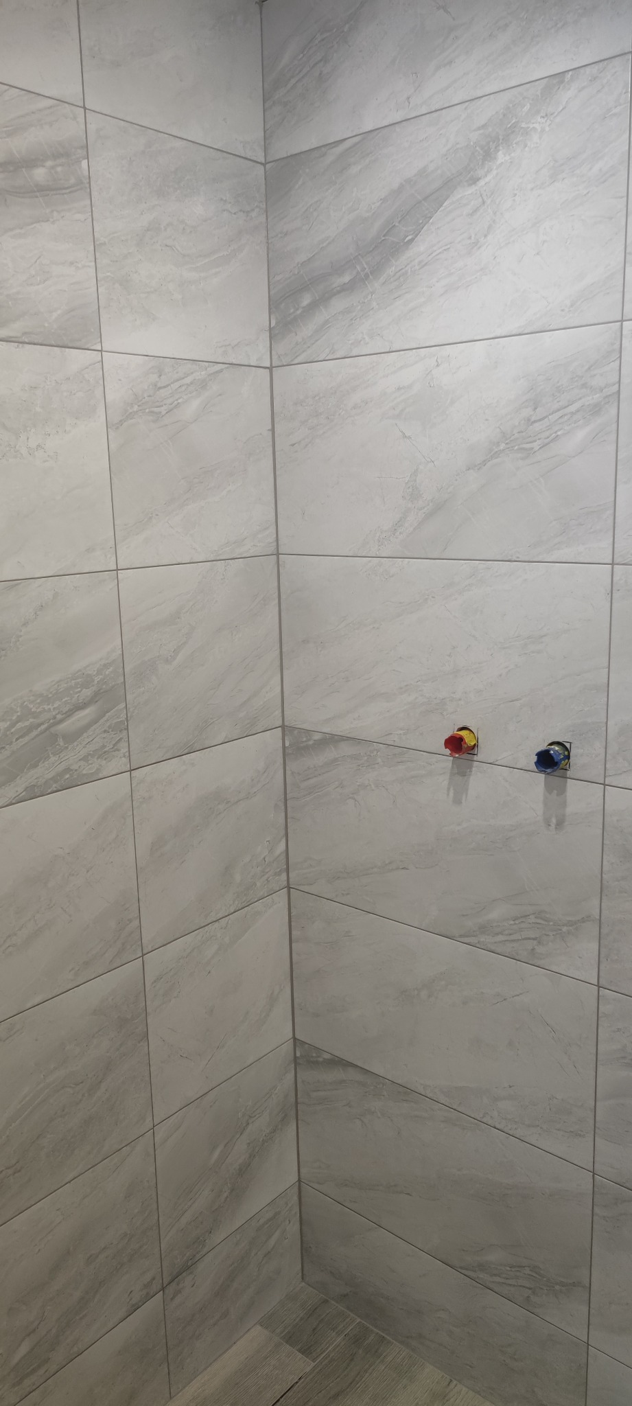 Wykończona ściana w łazience z szarymi płytkami imitującymi marmur oraz widocznymi przyłączami wodnymi z kolorowymi zaślepkami.