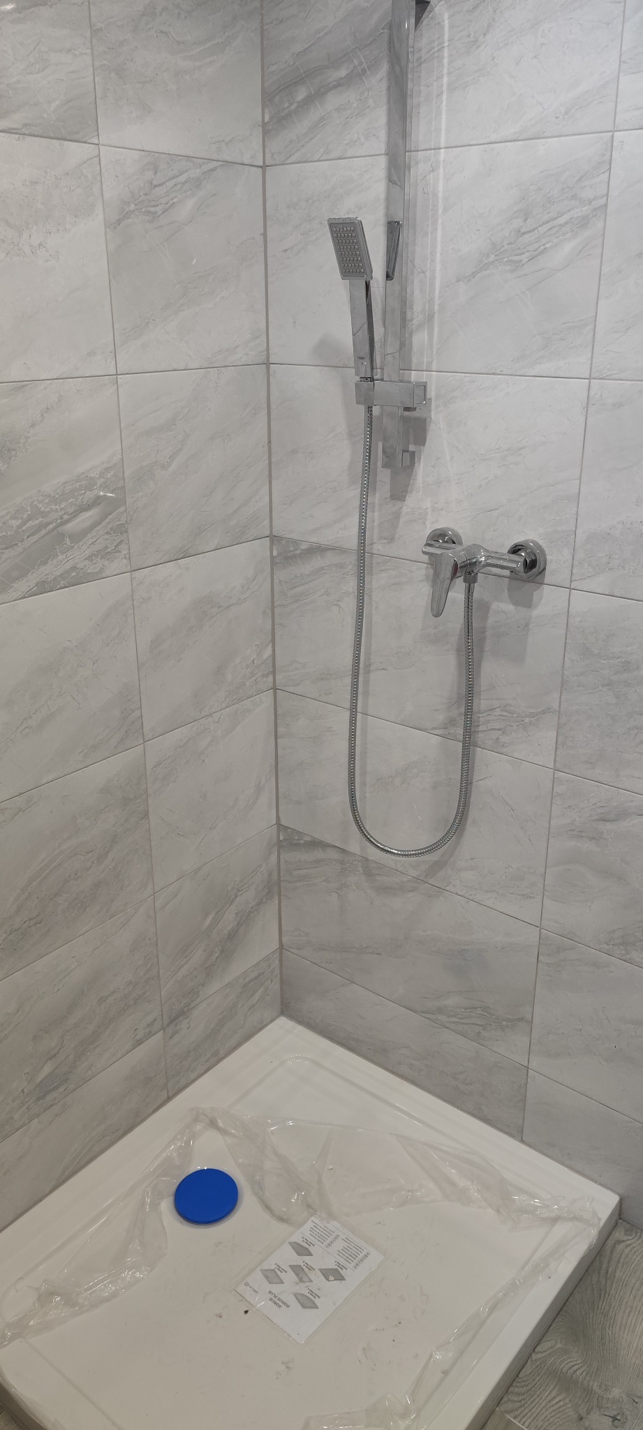 Wykończona kabina prysznicowa z szarymi, marmurkowymi płytkami ściennymi, chromowaną armaturą i brodzikiem zabezpieczonym folią ochronną z niebieskim korkiem odpływu.