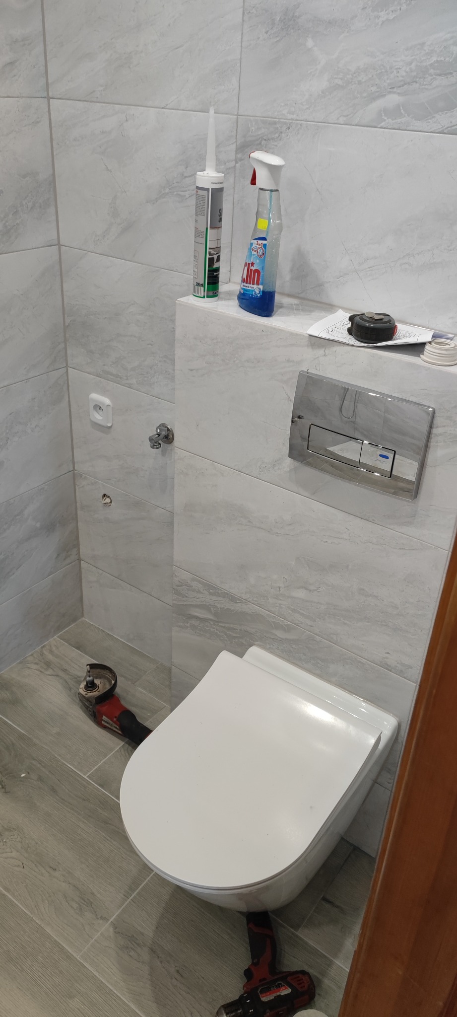 Nowoczesna toaleta wisząca z szarą płytką imitującą marmur na ścianach, widoczne narzędzia: szlifierka kątowa i wiertarka, a także silikon i płyn do czyszczenia, sugerujące trwający montaż.