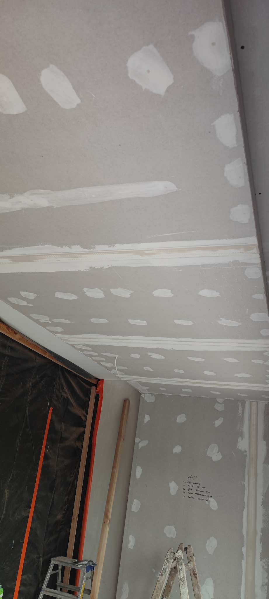 Wnętrze w trakcie prac wykończeniowych: sufit i ściany z płyt kartonowo-gipsowych, widoczne spoiny i miejsca po wkrętach zaszpachlowane, drabina oparta o ścianę, notatki na ścianie. Ochrona folią...