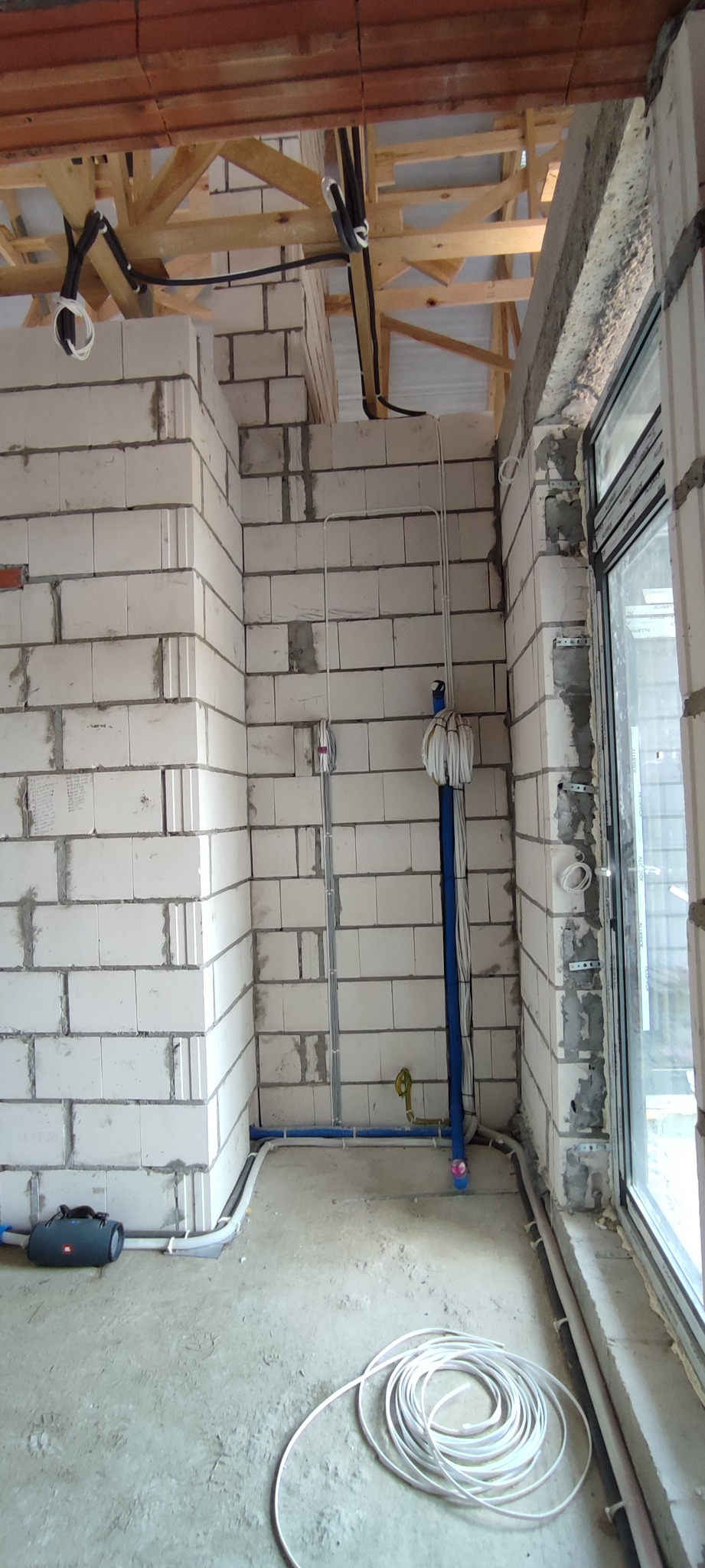 Surowy stan instalacji elektrycznej w pomieszczeniu z nieotynkowanymi ścianami z betonu komórkowego, widoczne kable prowadzone po ścianach i pod sufitem na drewnianej konstrukcji, zwój białego...