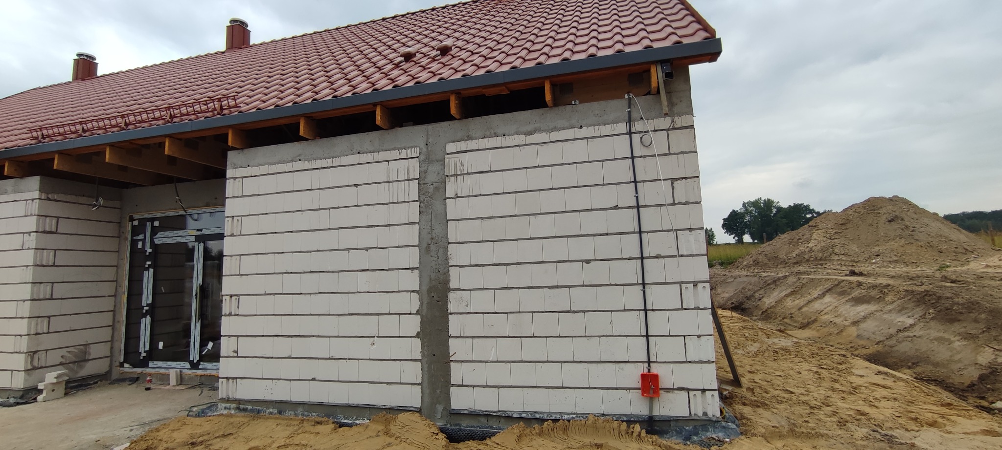 Niedokończony budynek jednorodzinny z białego bloczka, widoczna instalacja elektryczna w peszlu na ścianie zewnętrznej, dach pokryty czerwoną dachówką.