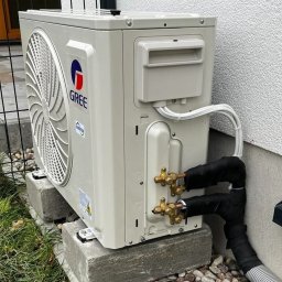 KOMFORT KLIMA JK ELWIRA KAMIŃSKA - Zewnętrzna jednostka klimatyzacji Gree zamontowana na betonowych podstawach przy ścianie budynku, z widocznymi rurami miedzianymi owiniętymi czarną taśmą izolacyjną i przewodami elektrycznymi.