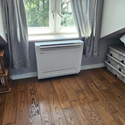 KOMFORT KLIMA JK ELWIRA KAMIŃSKA - Biały klimatyzator pod oknem, zasłoniętym szarymi zasłonami, na drewnianej podłodze. Obok regał z książkami i pojemniki z pokrywkami. Ustawiony w rogu pokoju.