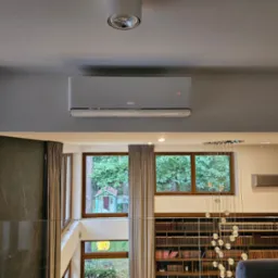 Szary klimatyzator marki Gree zamontowany na ścianie w nowoczesnym wnętrzu z widokiem na zieleń przez okna. Widoczna biblioteczka i żyrandol.