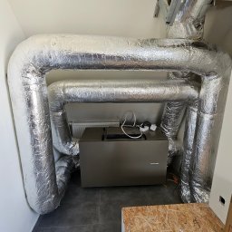 KOMFORT KLIMA JK ELWIRA KAMIŃSKA - Szare urządzenie Thessla Green otoczone izolowanymi, srebrnymi rurami wentylacyjnymi w białym pomieszczeniu. Widoczne kable i fragment podłogi.