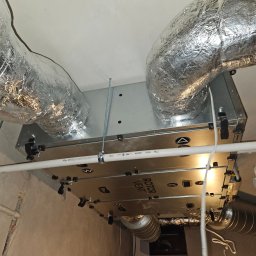 KOMFORT KLIMA JK ELWIRA KAMIŃSKA - Zamontowana centrala wentylacyjna Rotor Vent z izolowanymi przewodami w pomieszczeniu, widok z dołu na sufit. Elementy instalacji widoczne w kadrze.
