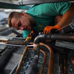 KOMFORT KLIMA JK ELWIRA KAMIŃSKA - Instalator w trakcie lutowania rur miedzianych, z użyciem palnika gazowego. Widoczne iskry, rękawice ochronne i elementy instalacji.