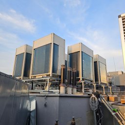 KOMFORT KLIMA JK ELWIRA KAMIŃSKA - Szereg agregatów klimatyzacyjnych na dachu budynku, widok z dołu, z butlami technicznymi w tle. Błękitne niebo z delikatnymi chmurami w górnej części kadru.
