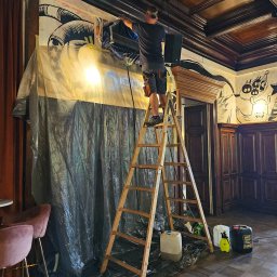 KOMFORT KLIMA JK ELWIRA KAMIŃSKA - Montaż klimatyzacji w lokalu: mężczyzna na drabinie instaluje urządzenie, zabezpieczone folią, w pomieszczeniu z dekoracyjnymi panelami ściennymi i wzorzystym sufitem.