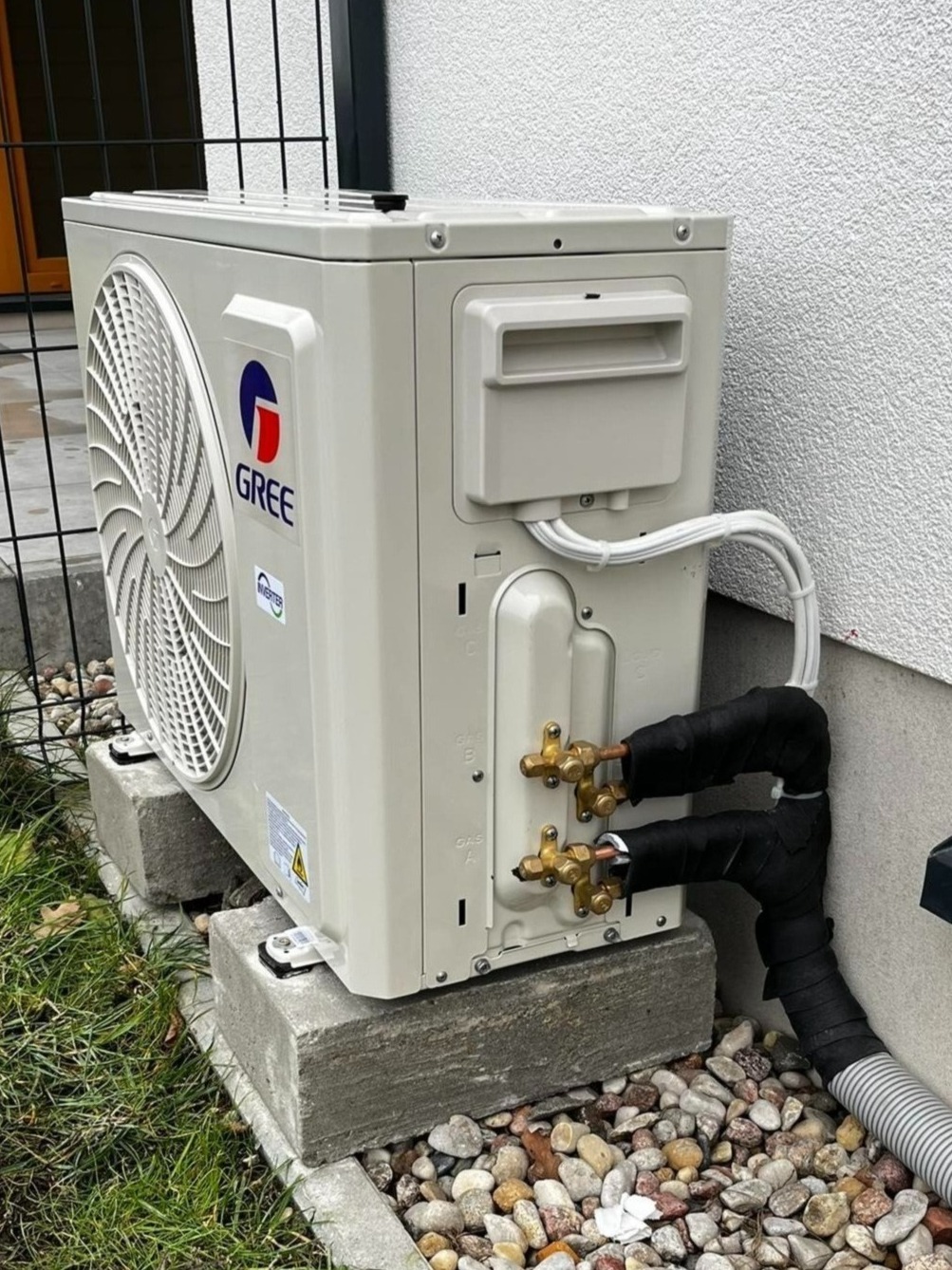 Zewnętrzna jednostka klimatyzacji Gree zamontowana na betonowych podstawach przy ścianie budynku, z widocznymi rurami miedzianymi owiniętymi czarną taśmą izolacyjną i przewodami elektrycznymi.