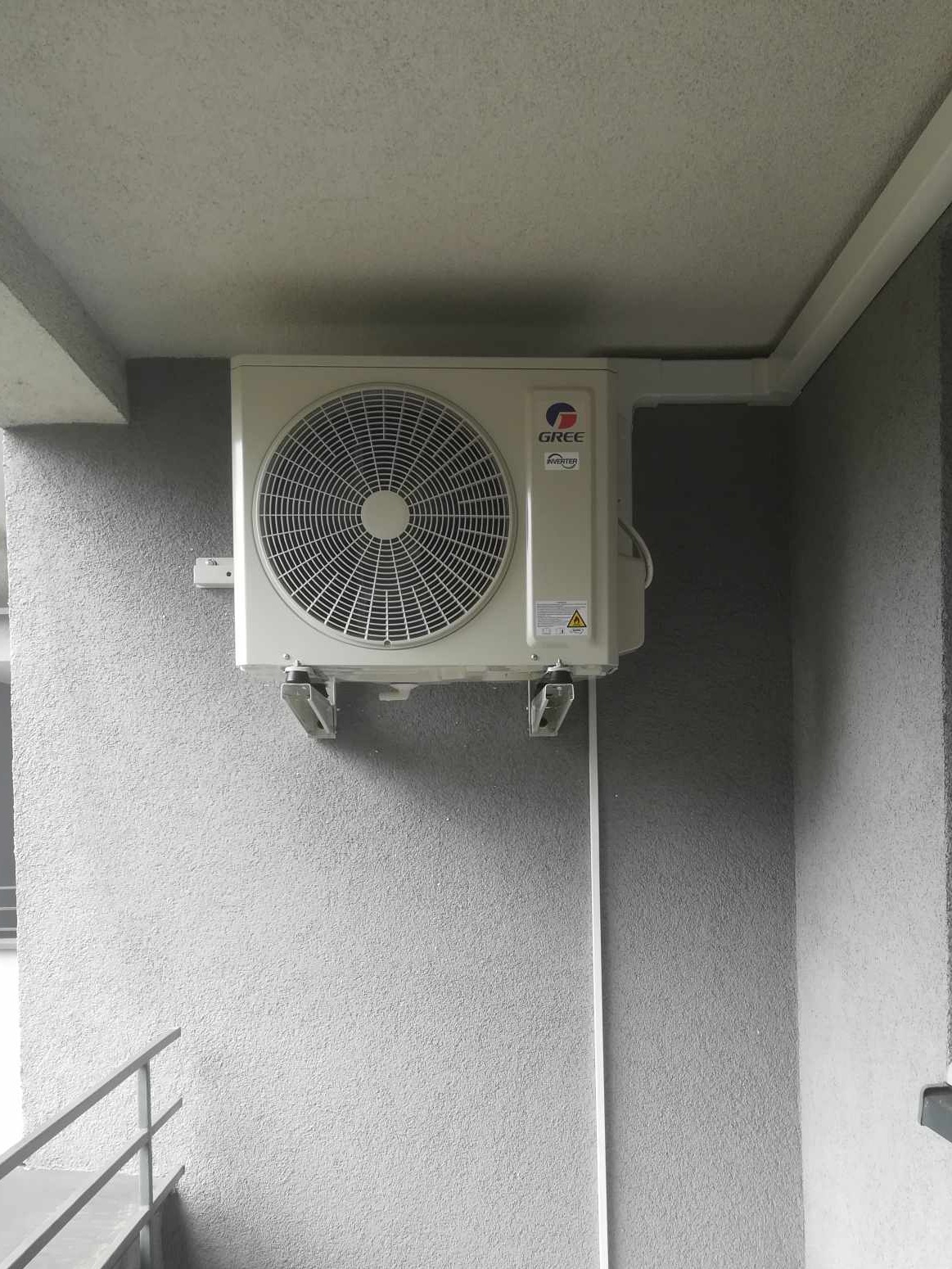 Zewnętrzna jednostka klimatyzacji marki Gree zamontowana na szarej, teksturowanej ścianie budynku, widoczne metalowe wsporniki i plastikowa listwa maskująca przewody.