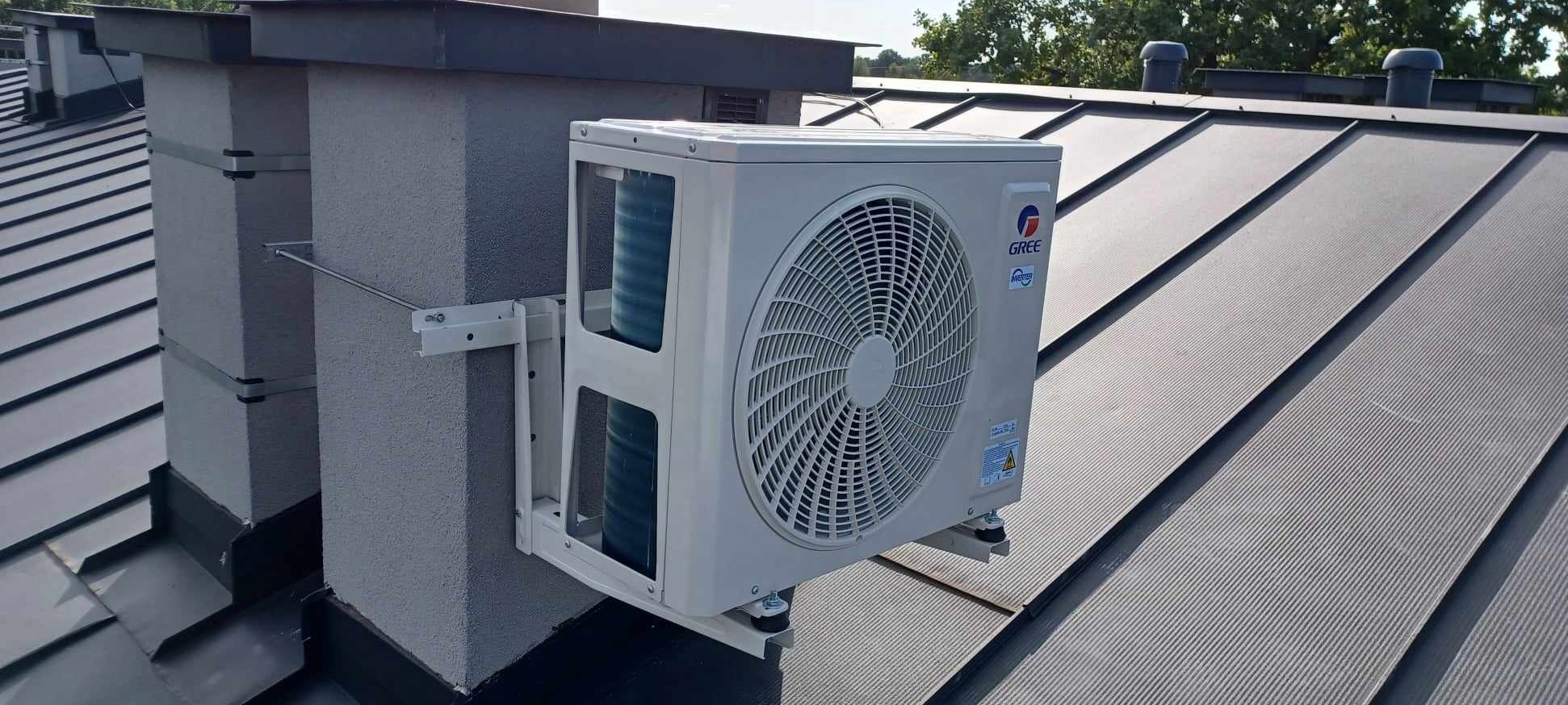 Zewnętrzna jednostka klimatyzacji marki Gree zamontowana na wspornikach przy kominie na dachu pokrytym blachą trapezową.
