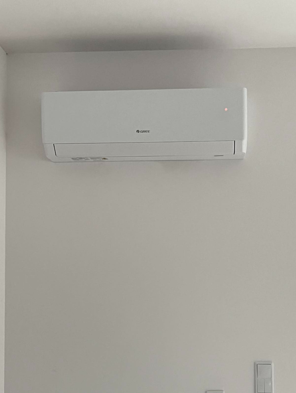 Biały klimatyzator marki Gree zamontowany na jasnej ścianie w pomieszczeniu mieszkalnym, widoczna delikatna faktura ściany i subtelne oświetlenie.