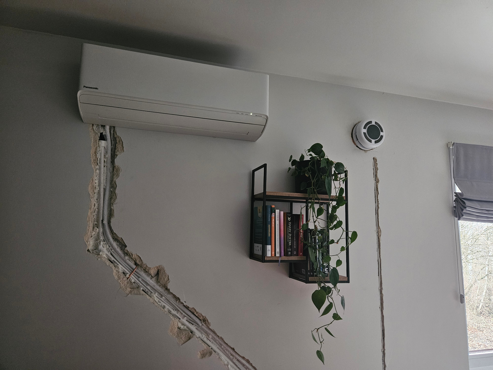 Biały klimatyzator Panasonic na ścianie z wyciętym rowkiem na kable. Obok półka z książkami i rośliną, czujnik dymu. Widoczne przewody elektryczne.