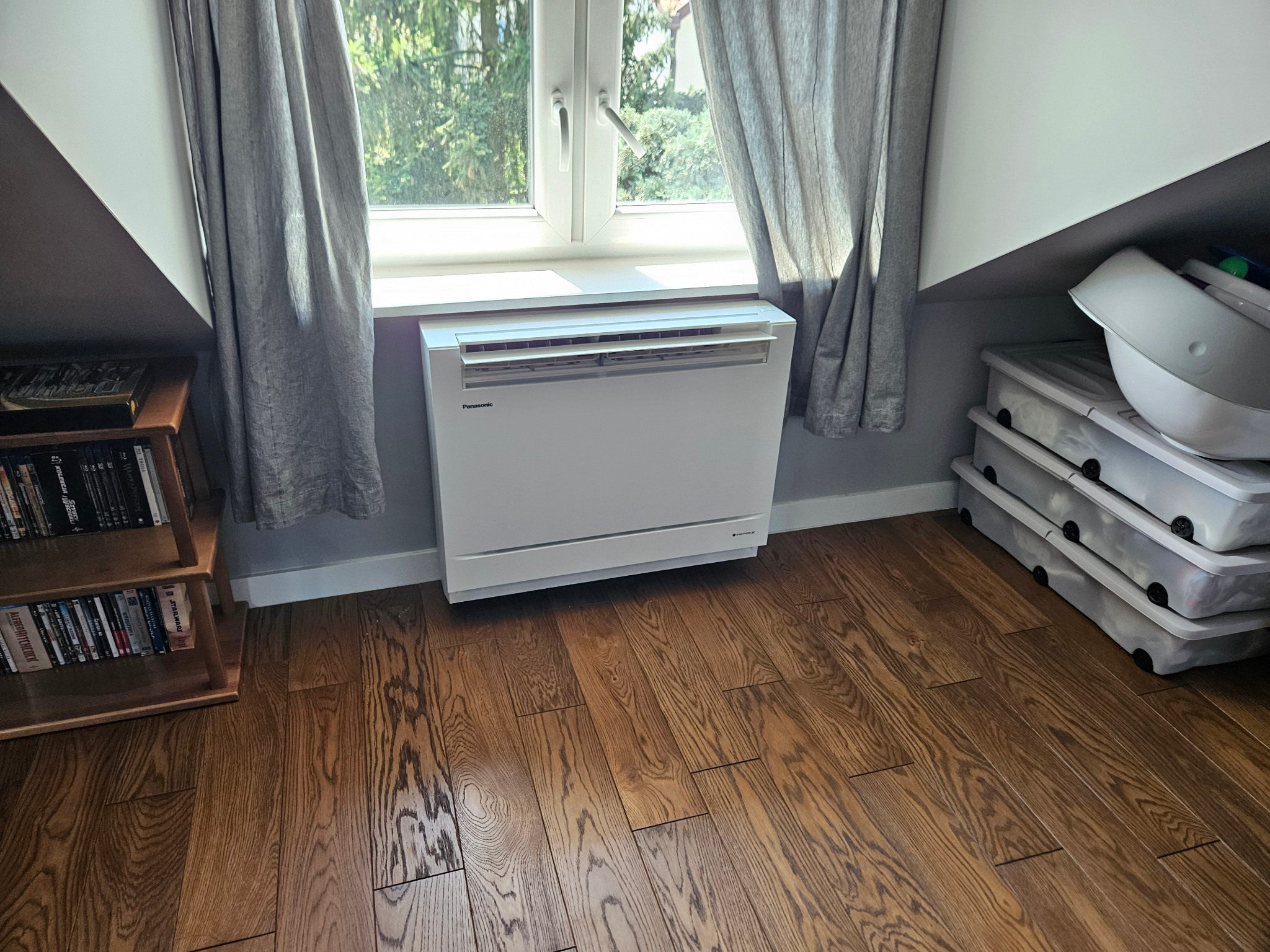 Biały klimatyzator pod oknem, zasłoniętym szarymi zasłonami, na drewnianej podłodze. Obok regał z książkami i pojemniki z pokrywkami. Ustawiony w rogu pokoju.