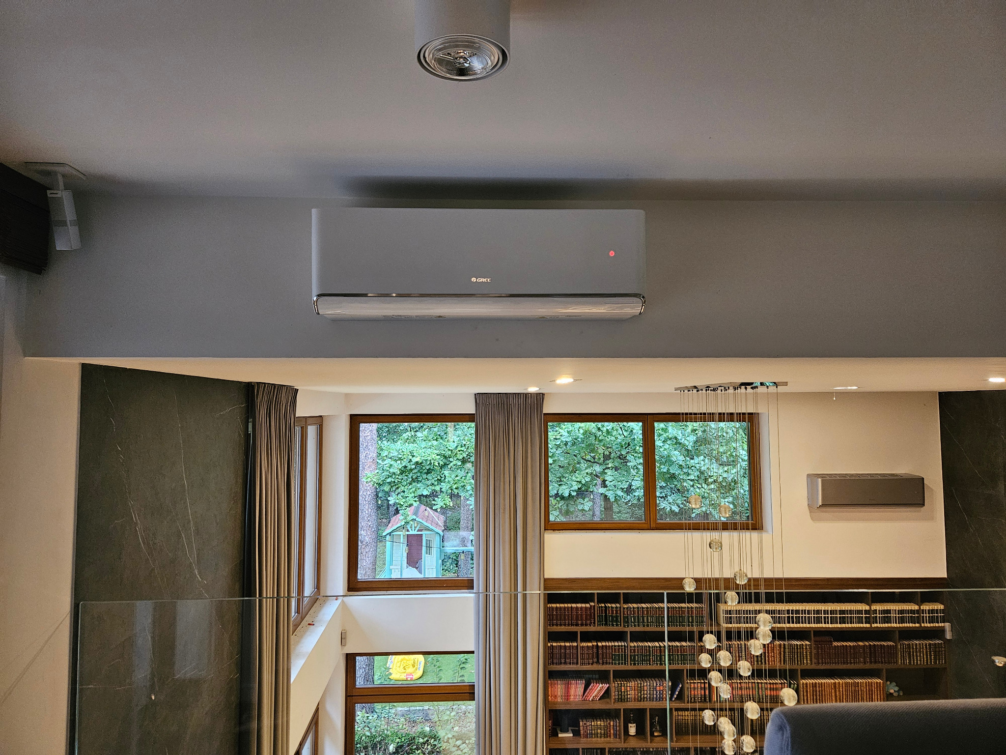 Szary klimatyzator marki Gree zamontowany na ścianie w nowoczesnym wnętrzu z widokiem na zieleń przez okna. Widoczna biblioteczka i żyrandol.