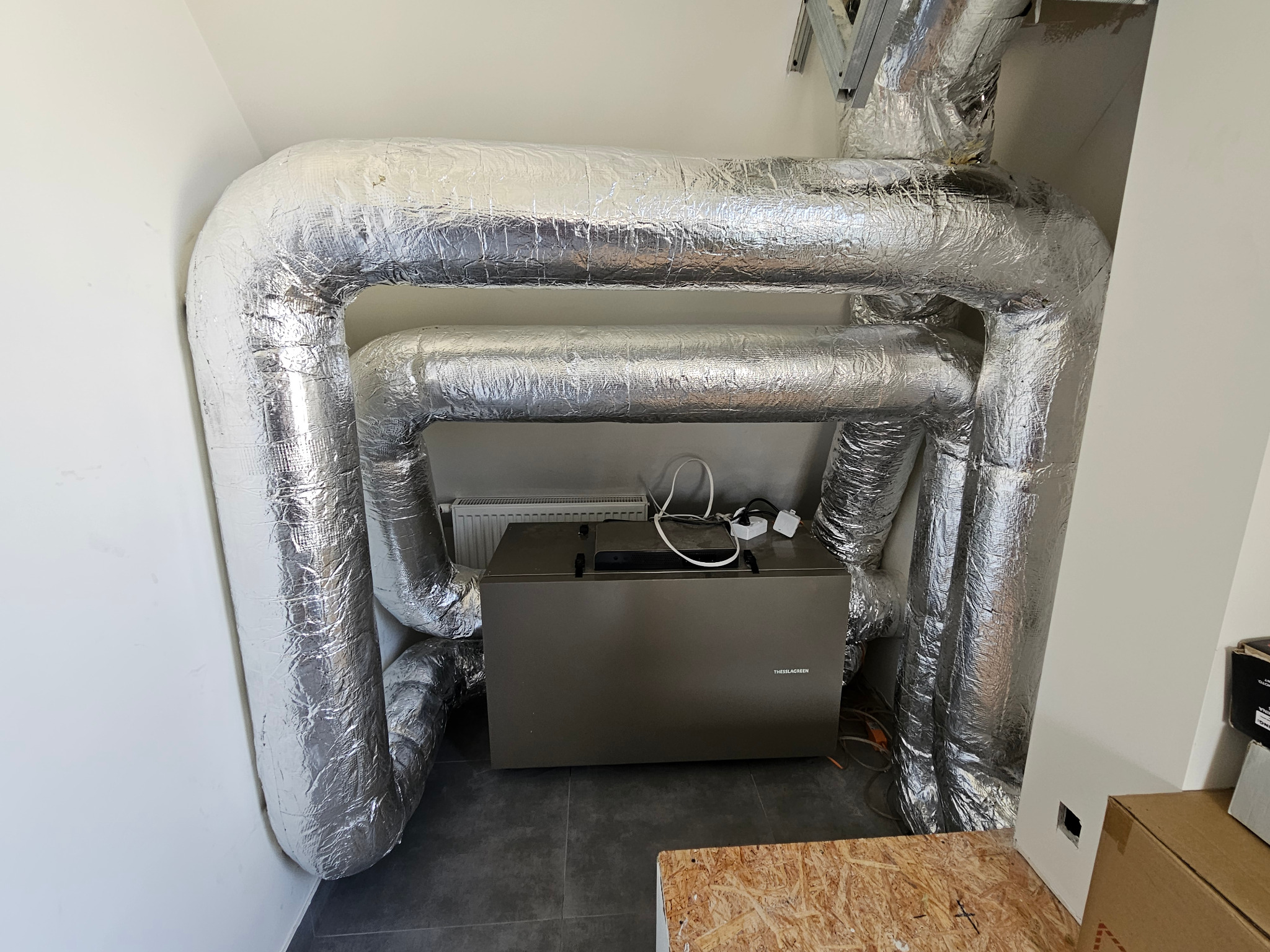 Szare urządzenie Thessla Green otoczone izolowanymi, srebrnymi rurami wentylacyjnymi w białym pomieszczeniu. Widoczne kable i fragment podłogi.
