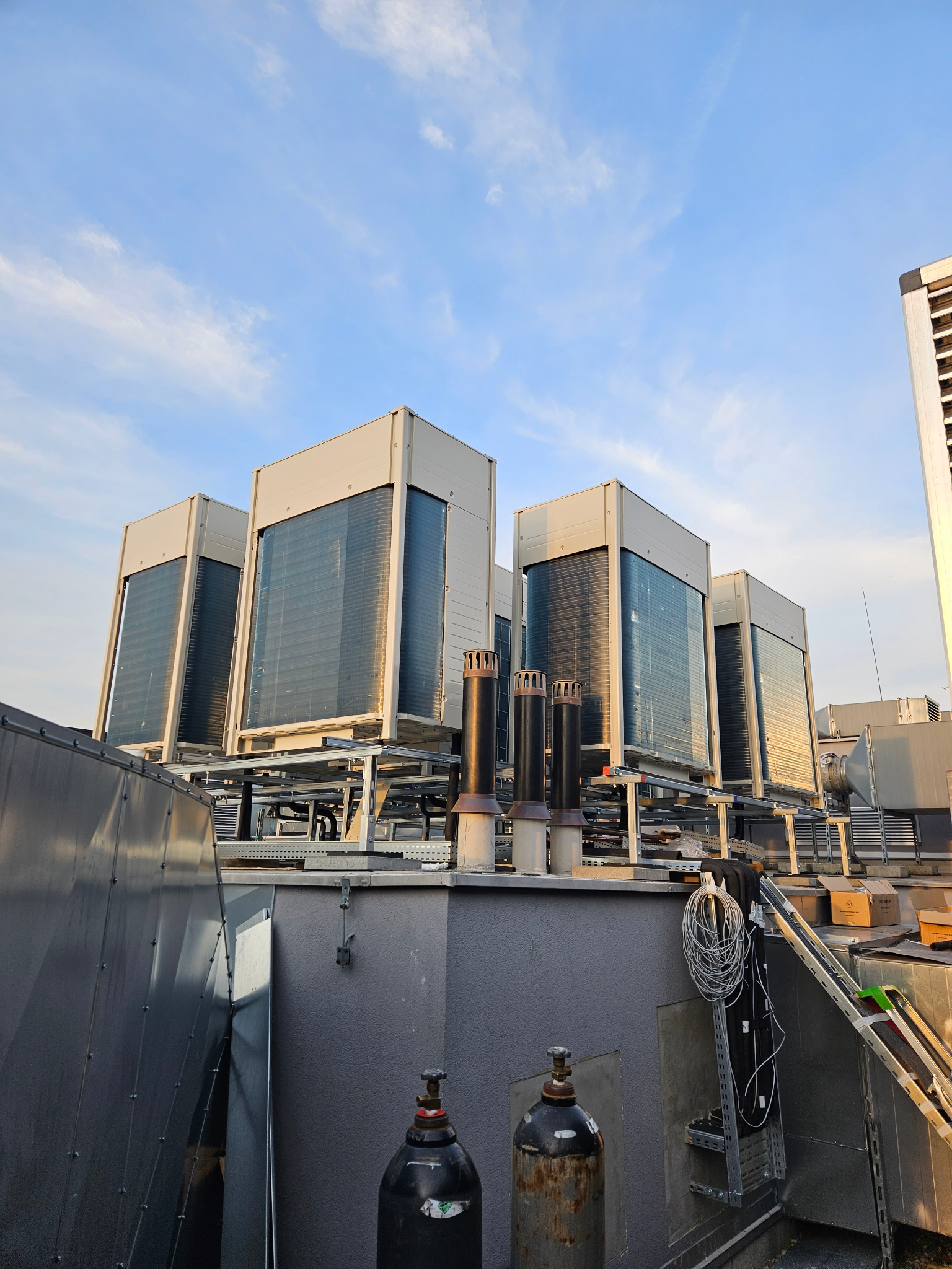 Szereg agregatów klimatyzacyjnych na dachu budynku, widok z dołu, z butlami technicznymi w tle. Błękitne niebo z delikatnymi chmurami w górnej części kadru.