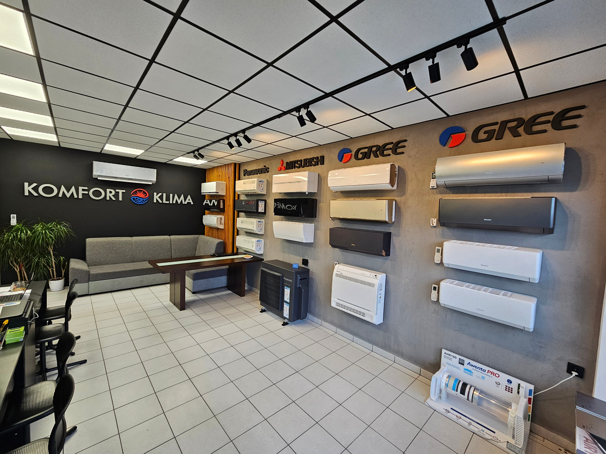 Salon klimatyzacji z ekspozycją modeli Panasonic, Mitsubishi i Gree. Widoczne sofy, biurka i regały z urządzeniami. Oświetlenie sufitowe punktowe i kasetonowe.
