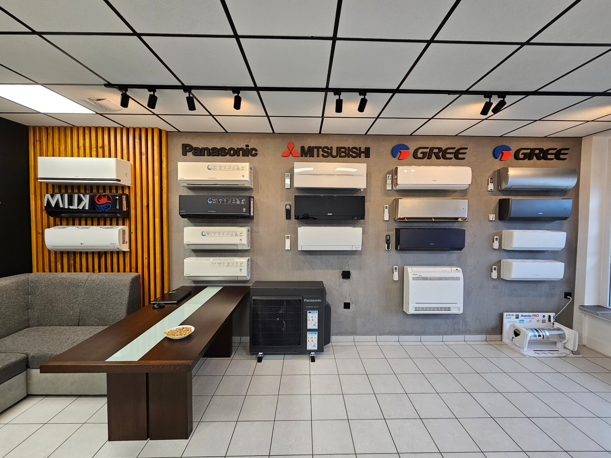 Salon z ekspozycją klimatyzatorów różnych marek (Panasonic, Mitsubishi, Gree) na ścianie. Widoczny model podłogowy Panasonic, stół z przekąskami i szara sofa.