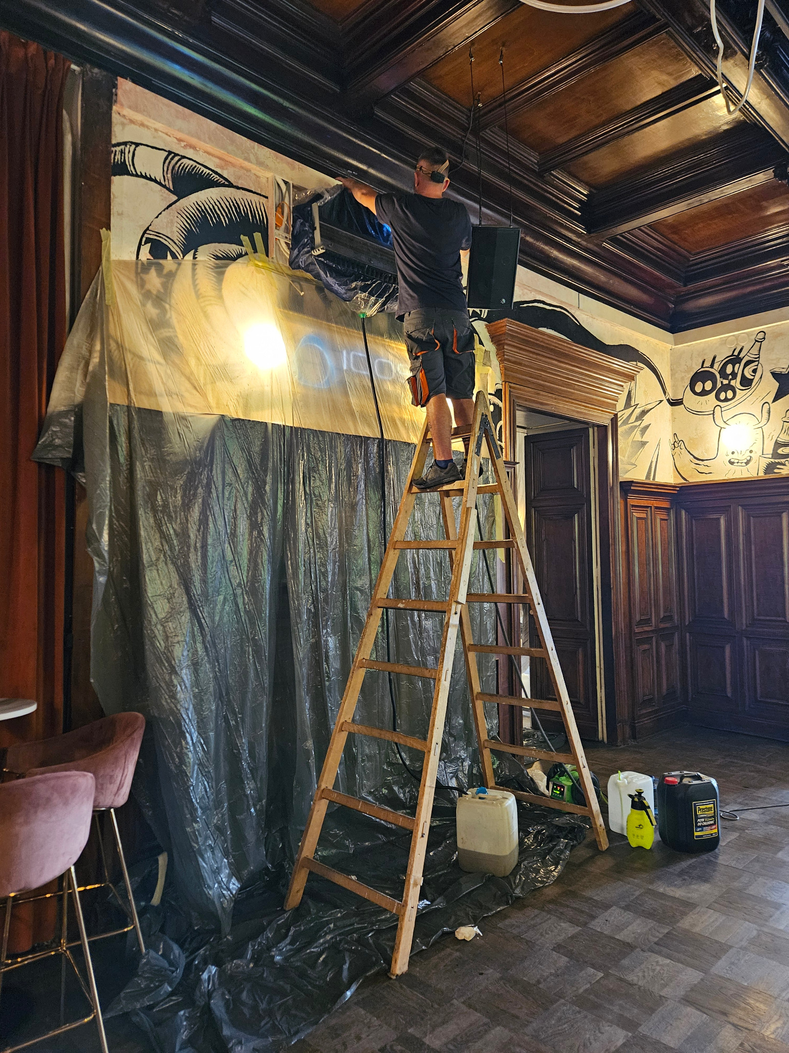 Montaż klimatyzacji w lokalu: mężczyzna na drabinie instaluje urządzenie, zabezpieczone folią, w pomieszczeniu z dekoracyjnymi panelami ściennymi i wzorzystym sufitem.