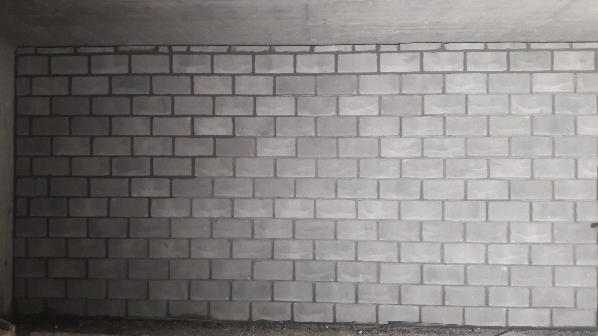 Świeżo wymurowana ściana z szarych bloczków betonowych, widoczna spod sufitu, z ciemniejszymi spoinami i drobnymi resztkami zaprawy na podłodze.