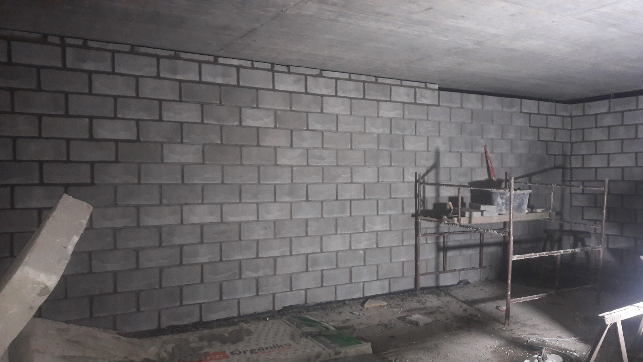 Świeżo wymurowana ściana z szarych bloczków betonowych w surowym wnętrzu, widoczne rusztowanie z narzędziami i resztkami zaprawy, betonowy sufit.