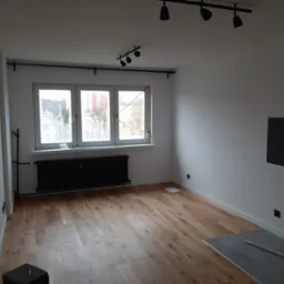 Wykończony pokój z parkietem w jodełkę, białymi ścianami, czarnym grzejnikiem i oświetleniem punktowym na suficie, widok z niskiego kąta.