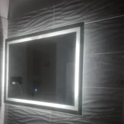 Podświetlane lustro w łazience z szarymi płytkami o falistej strukturze, widoczny kontakt z ładowarką i dotykowy włącznik.