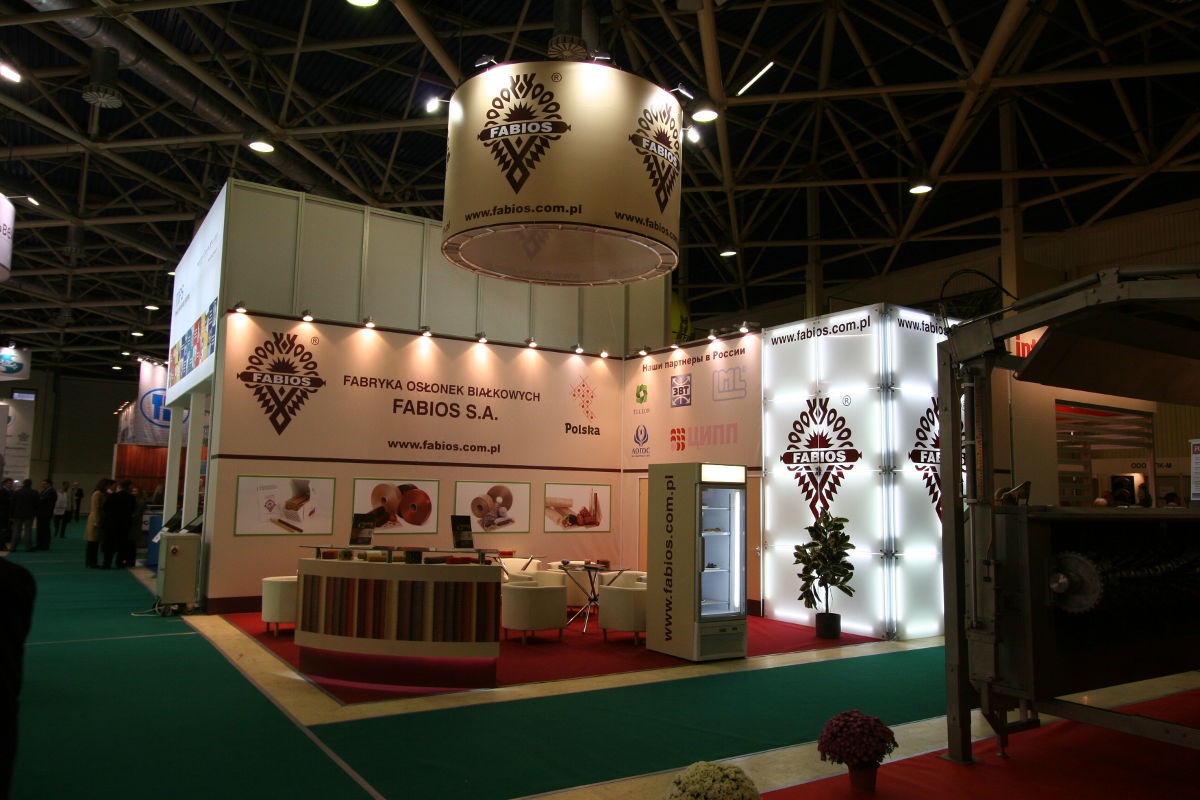 Stoisko targowe z podwieszonym cylindrem reklamowym, prezentujące firmę produkującą osłonki białkowe, z oświetlonymi ścianami i ekspozycją produktów.