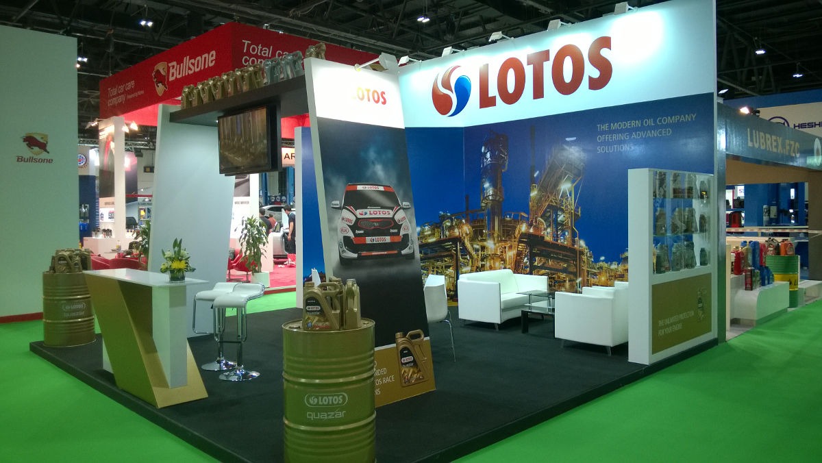 Stoisko targowe z logo LOTOS, prezentujące samochód wyścigowy i produkty Quazar na tle rafinerii, z białymi sofami i barem.