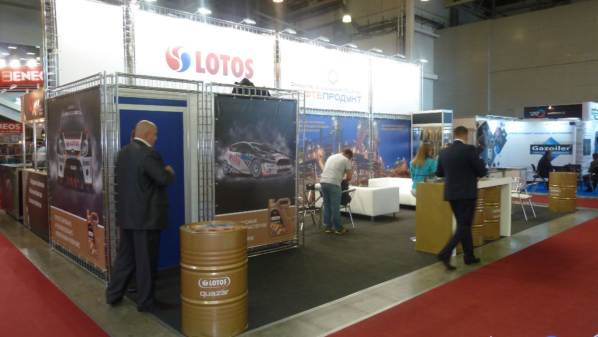 Stoisko targowe z logo Lotos, prezentujące oleje silnikowe Quazar w beczkach i na grafikach, z pracownikami w tle.