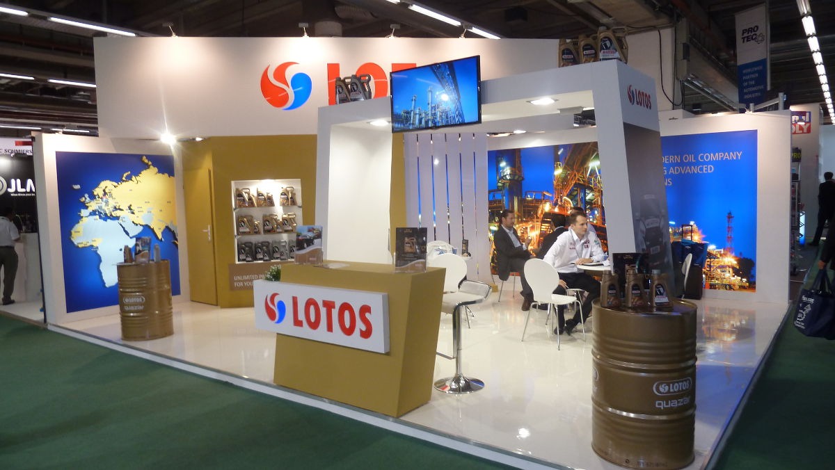 Stoisko targowe firmy Lotos z ekspozycją produktów, dużym logo, ekranem wyświetlającym rafinerię i miejscami do rozmów z klientami.