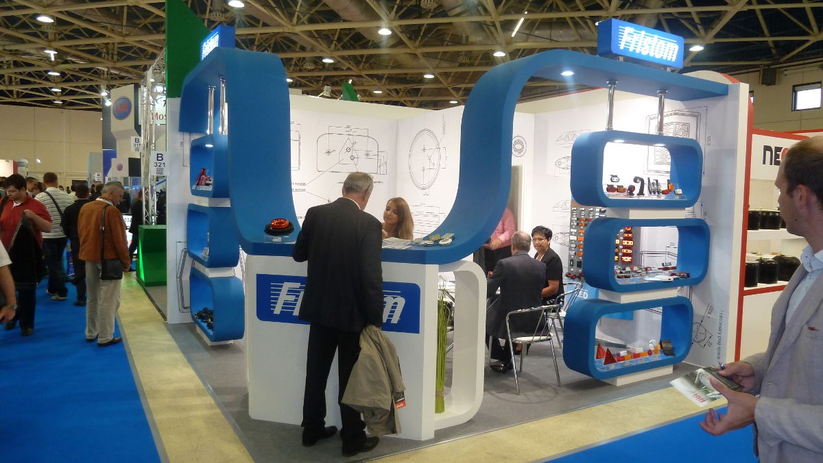 Stoisko targowe firmy z niebieskimi elementami konstrukcyjnymi i logo Fristom, prezentujące oświetlenie samochodowe i akcesoria, otoczone przez zwiedzających.