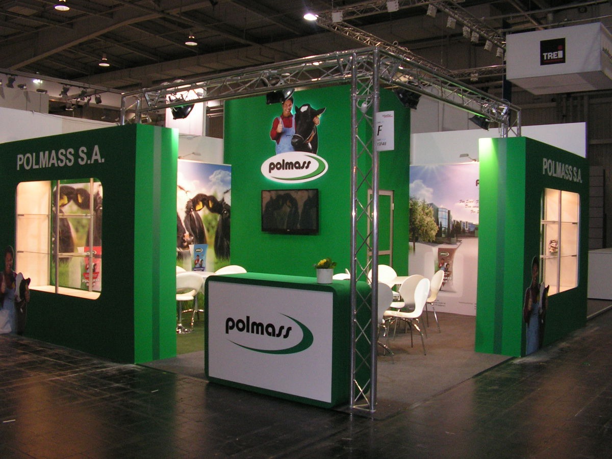 Stoisko targowe firmy Polmass S.A. z zielonymi ścianami, grafikami krów i metalową konstrukcją sufitową oświetloną reflektorami.