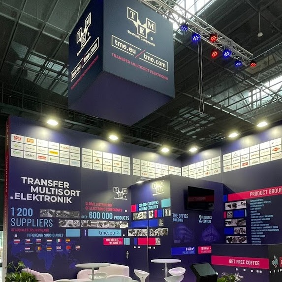Stoisko targowe z podwieszonym, sześciennym banerem z logo TME, oświetlone reflektorami, z ekspozycją produktów i informacjami o firmie na ścianach.