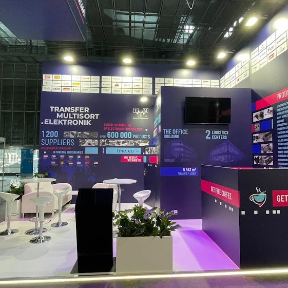 Stoisko targowe firmy TME (Transfer Multisort Elektronik) z logo, informacjami o firmie, miejscami do siedzenia i roślinami doniczkowymi.